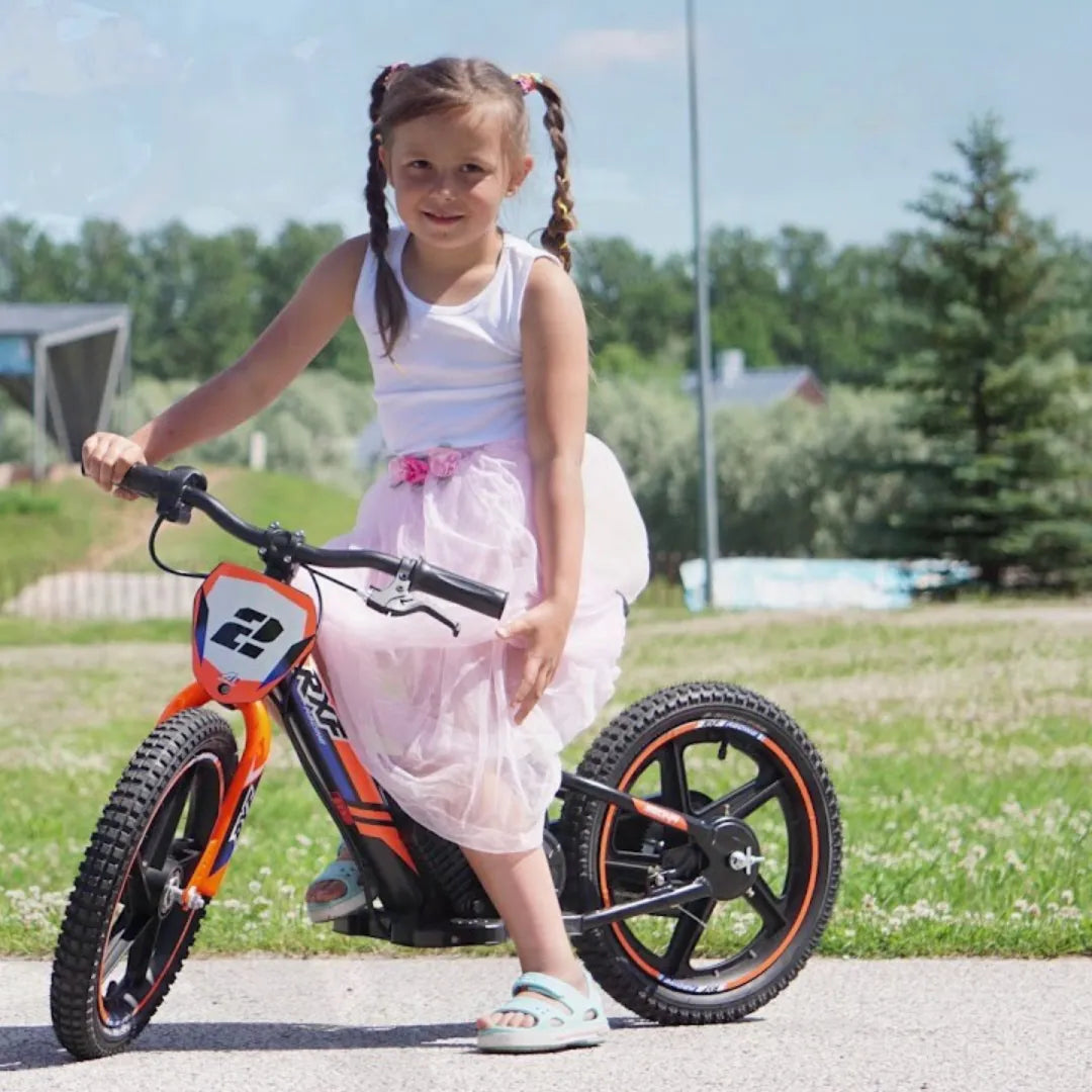 Izdelek na sliki je električno kolo, model Apollo Sedna 12 RXF. Na sliki je prikazana deklica, ki sedi na kolesu, oblečena v belo majico in roza krilo. Kolo ima svetlo oranžno barvo z značilnim dizajnom in širokimi gumami, kar daje videz športnega in otroškega kolesa. V ozadju je zeleno okolje, kar nakazuje, da je slika posneta na prostem.