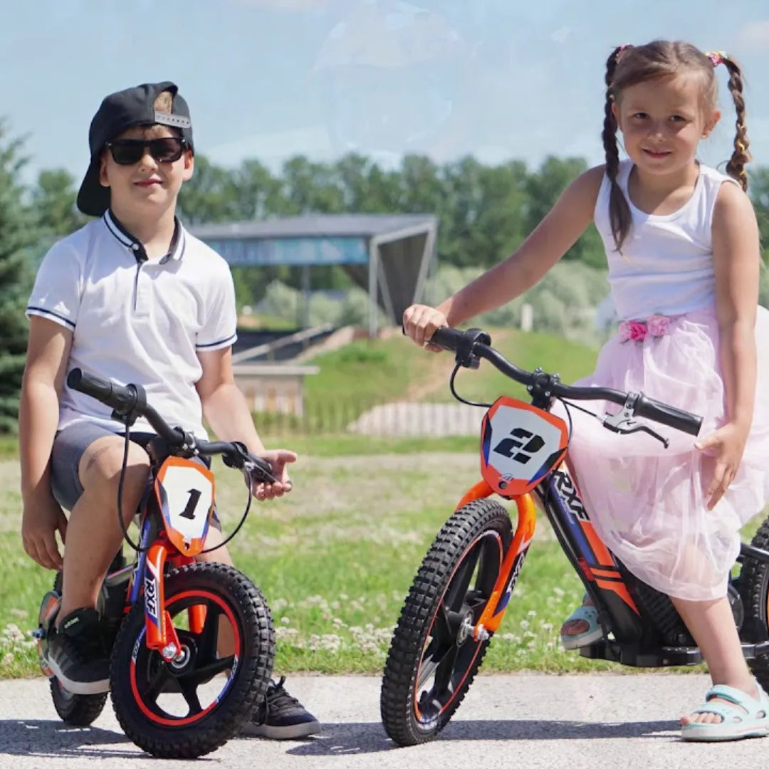 Izdelek na sliki je otroško električno kolo znamke RXF, model Apollo Sedna 16. Na sliki sta dva otroka, deček in deklica, ki sedita na kolesih. Deček nosi sončna očala in kapo, deklica pa oblečeno belo majico in roza krilo. Obe kolesi sta živahne oranžne barve z oznakami številk na sprednjem delu. V ozadju je zeleno okolje s športnim objektom.