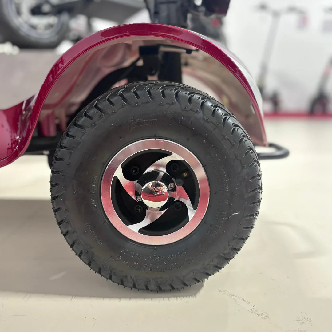 Izdelek je električni mobilni voziček, model Eco Speed Senior. Na sliki je vidna zadnja pnevmatika vozička, ki ima robusten profil in aluminijasto platišče. Barva vozička je rdeča, kar daje izdelku privlačen videz. V ozadju so vidni tudi drugi električni skiroji, kar nakazuje, da gre za prodajno okolje.