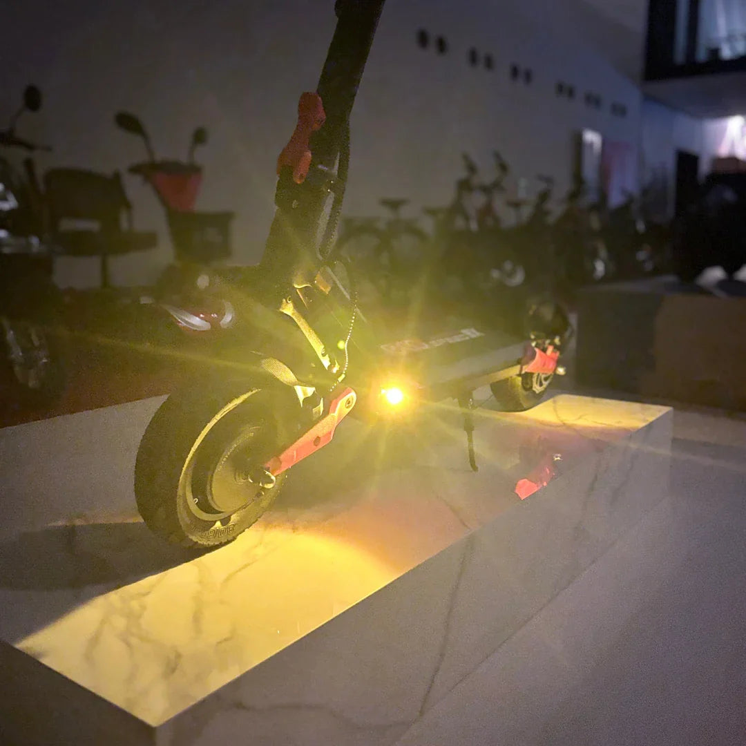 Na sliki je prikazan električni skiro model Eco Speed 9X. Vidna je zadnja kolesa skiroja, ki ima na sebi svetilko, ki sveti v rumeni barvi. Skira je postavljen na svetlo podlago, kar poudarja njegov dizajn in funkcionalnost. V ozadju so vidni tudi drugi skiroji, kar nakazuje, da gre za prostor, kjer so shranjeni različni modeli.