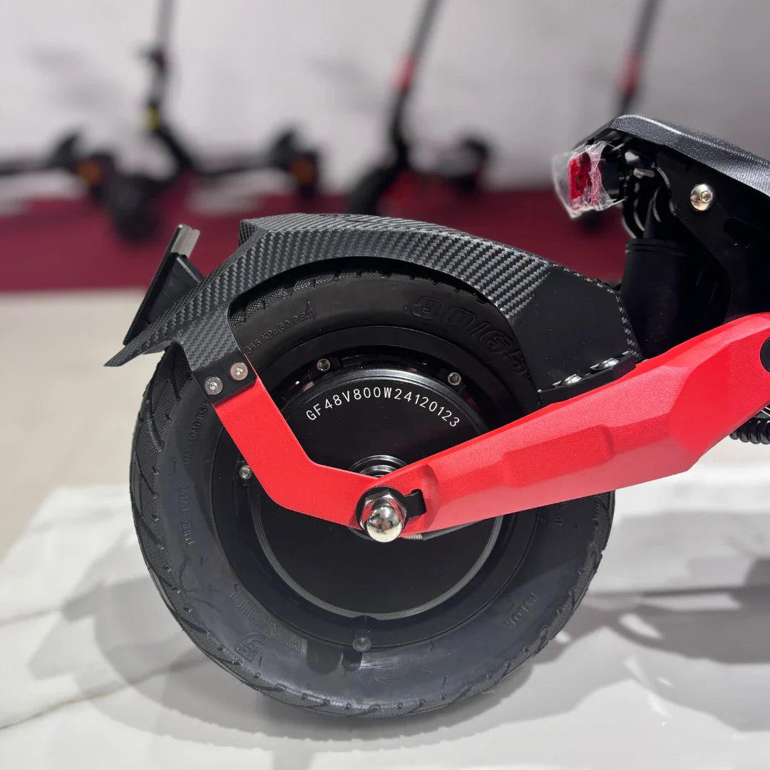 Na sliki je prikazan električni skiro model Eco Speed 9. Vidna je zadnja kolesna enota, ki vključuje motor in gumijasto pnevmatiko. Kolesna enota je opremljena s kovinsko rdečo obrobo in karbonsko zaščito, kar daje skiroju športen videz. V ozadju so vidni še drugi električni skiroji, kar nakazuje, da gre za razstavo ali prodajno mesto.