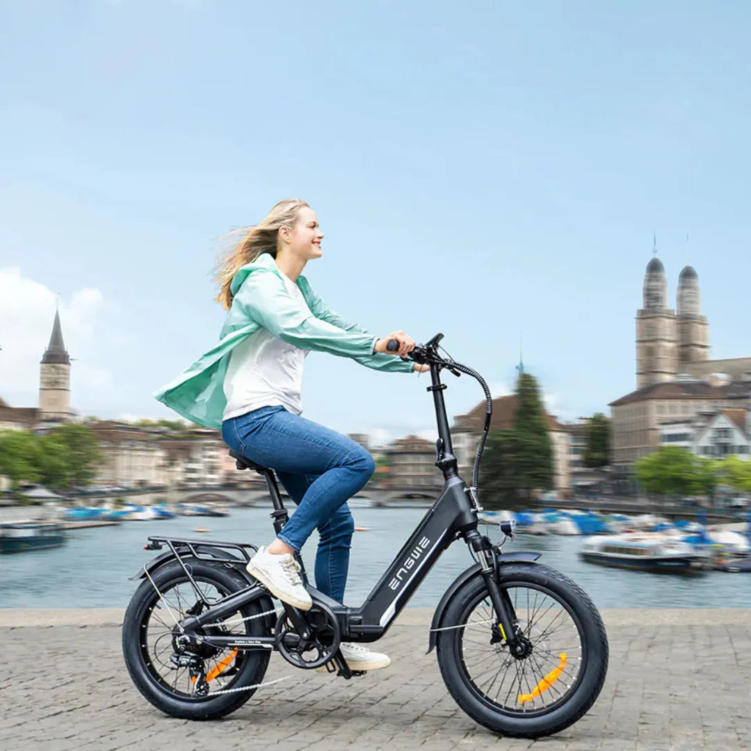 Izdelek na sliki je električno kolo znamke Engwe, model L20 3.0 Boost. Na sliki je prikazana mlada ženska, ki se vozi na kolesu ob vodi, v ozadju pa so vidne stavbe in ladje. Kolo ima kompaktno zasnovo, široke pnevmatike in je opremljeno s prednjimi in zadnjimi zavorami. Ženska nosi svetlo jakno in se smehlja, kar nakazuje užitek pri vožnji.