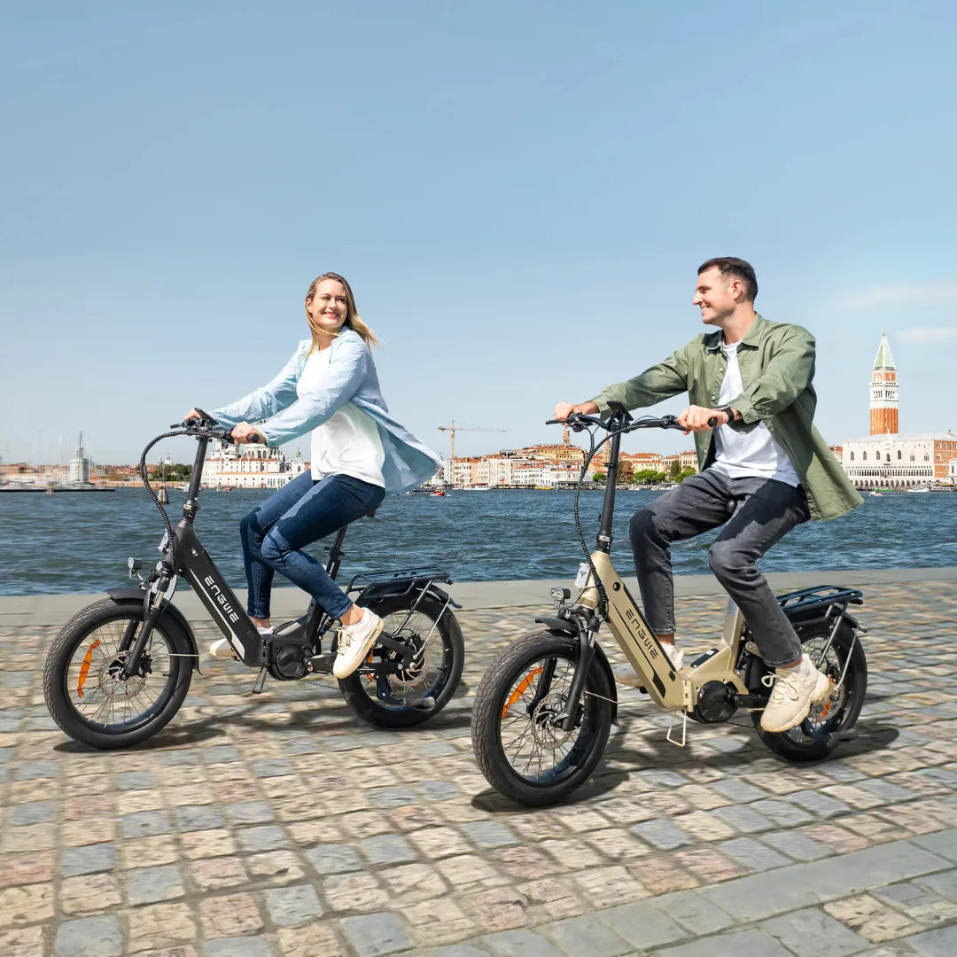 Na sliki sta prikazana dva električna kolesa znamke Engwe, model L20 3.0 Pro. Kolesa imata robustno zasnovo z debelimi pnevmatikami in električnim motorjem. Uporabnika se peljeta ob vodi, kar ustvarja sproščeno in prijetno vzdušje. Obe kolesi sta opremljeni s praktičnimi dodatki, kot so prtljažniki, kar jih naredi primerne za mestno vožnjo. V ozadju je vidna slikovita urbana pokrajina.