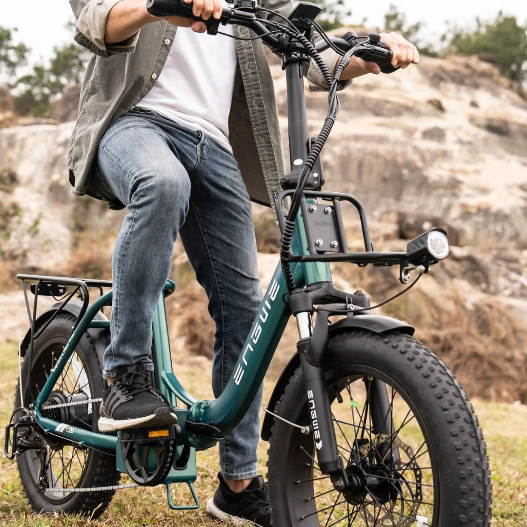 Na sliki je električno kolo znamke Engwe, model L20 Boost. Kolo ima široke pnevmatike, kar omogoča boljšo stabilnost in udobje pri vožnji po različnih terenih. Oseba na kolesu nosi športna oblačila in se nahaja na naravni lokaciji z skalnatimi površinami v ozadju. Kolo je opremljeno z nosilcem in svetilko, kar nakazuje na njegovo praktičnost za mestno in terensko uporabo.