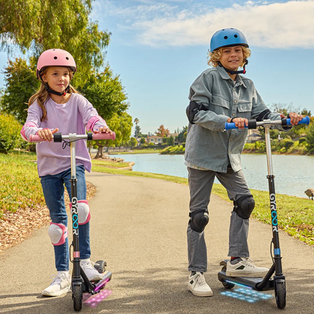 Izdelek je električni skiro, model Gyroor H30 Max. Na sliki sta vidna dva otroka, ki se vozita na skirojih ob obrežju, oblečena sta v zaščitno opremo, vključno s čeladami in zaščitnimi koleni. Okolica je zelena, ob vodi, kar ustvarja prijetno vzdušje za vožnjo.