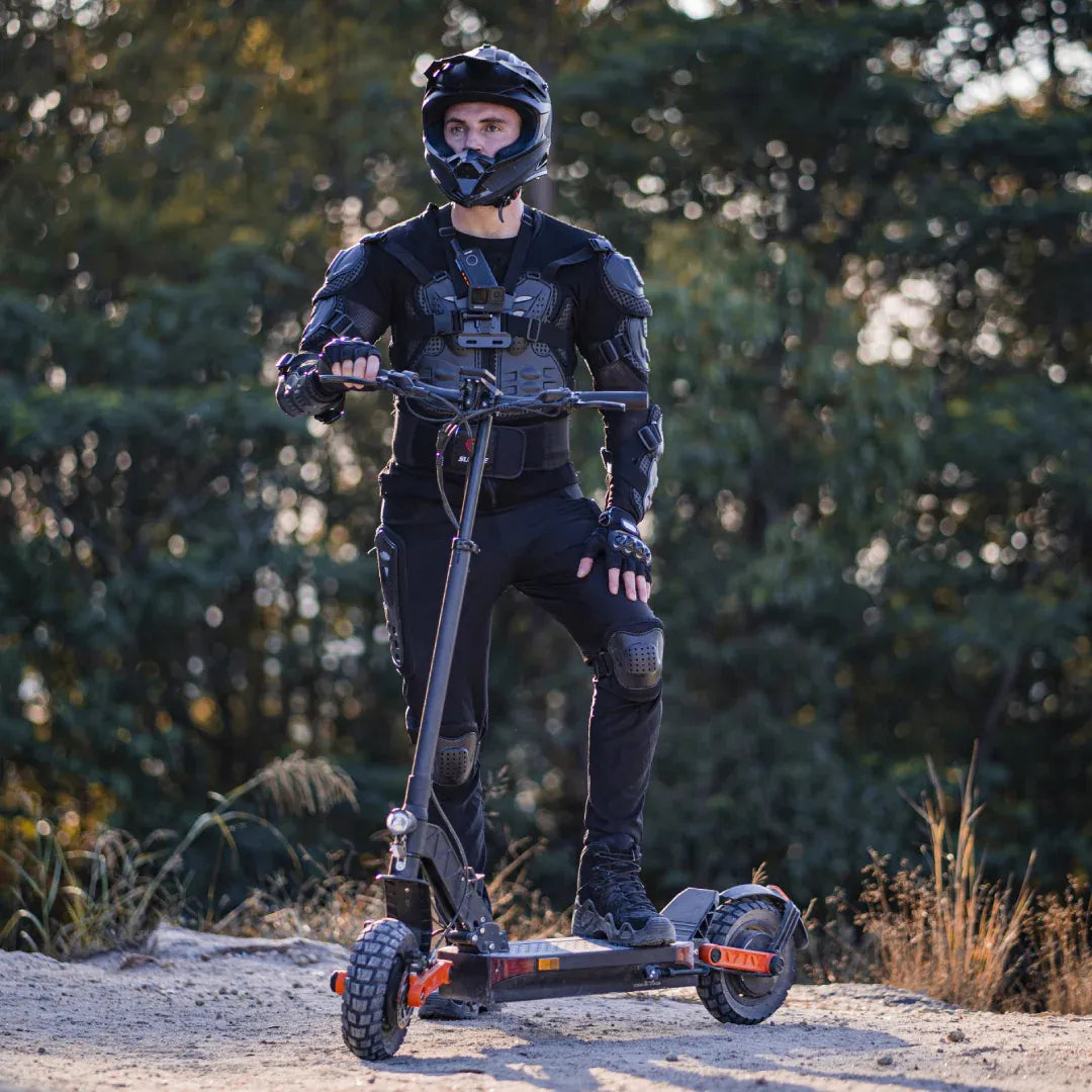 Izdelek na sliki je električni skiro, model Joyor S10 S-Z. Na sliki je moški, oblečen v črno zaščitno opremo in čelado, ki stoji na skiroju na zunanji površini. Okolica je naravna, z drevesi v ozadju. Scooterska zasnova je robustna, kar nakazuje na primernost za različne terene.