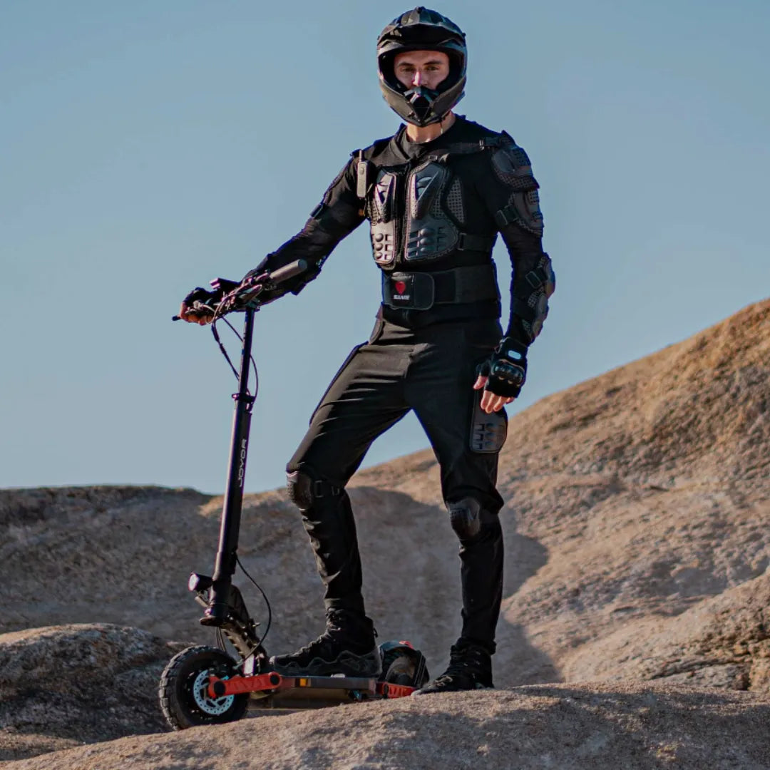 Na sliki je prikazan električni skiro, model Joyor S10 S-Z. Oseba, ki stoji na skiroju, nosi zaščitno opremo, vključno s čelado in zaščitnimi oblačili, kar kaže na to, da se pripravlja na vožnjo v zahtevnejših terenih. Skrbno izbrana lokacija na skalnatem terenu dodatno poudarja zmogljivosti skiroja.