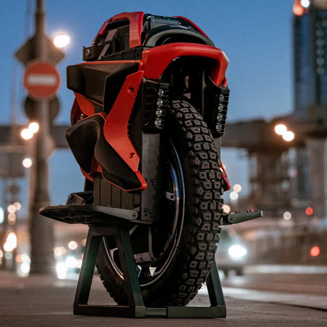 Izdelek na sliki je električni monocikel, model Kingsong S22 Pro. Na sliki je prikazan monocikel v rdeči in črni barvi, postavljen na stojalo. Vidna je velika guma z izrazitim profilom, kar nakazuje na dobro oprijemljivost, ter detajli na telesu monocikla, ki vključujejo oblikovane linije in LED luči. Ozadje prikazuje mestno okolje z uličnimi lučmi in prometnimi znaki.