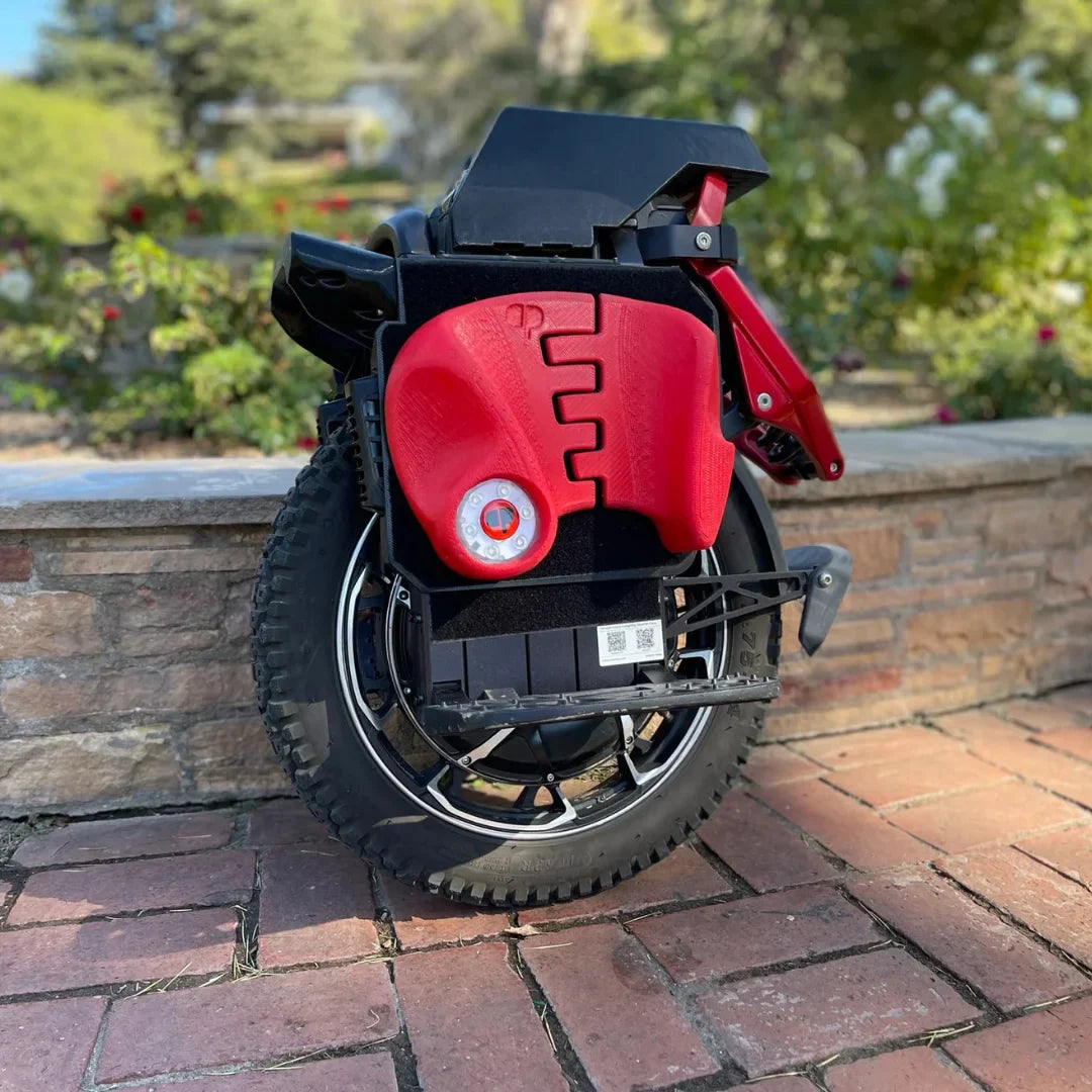 Izdelek na sliki je električni monocikel, model Kingsong S22 Pro. Na sliki je prikazan zložen monocikel, ki ima črno in rdečo barvo. Vidna je velika kolesa z robustnim profilom ter del, kjer se nahajajo kontrole in baterija. Monocikel je postavljen na kamenito površino, v ozadju pa so vidne rastline, kar daje občutek zunanjega okolja.