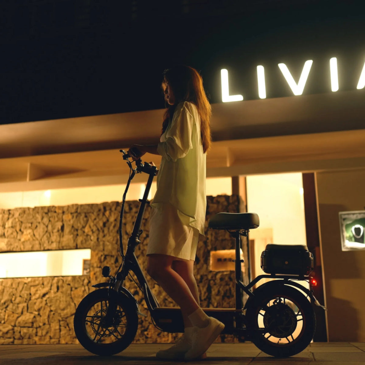 Izdelek na sliki je električni skiro, model Kukirin C1 Pro. Na sliki je vidna oseba, ki stoji ob skiroju, ki ima sedež in je zasnovan za udobno vožnjo. Osvetlitev v ozadju ustvarja prijetno nočno vzdušje, kar dodatno poudari modernost in funkcionalnost skiroja.