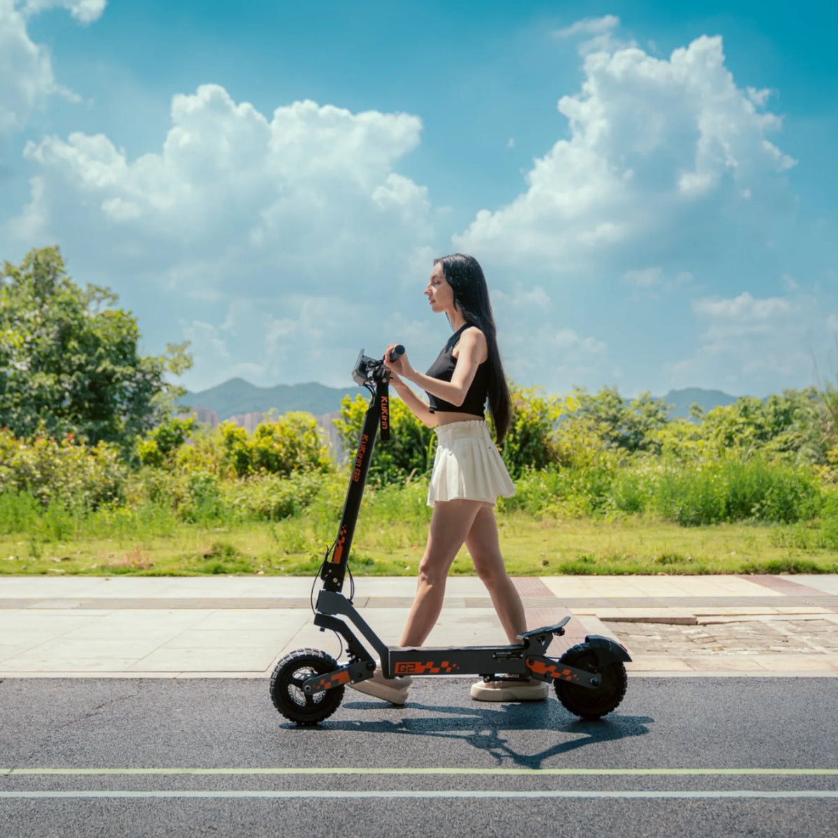 Na sliki je prikazan električni skiro znamke Kukirin, model G2. Vidimo mlado žensko, ki stoji na skiroju in se vozi po široki poti ob zelenem okolju. V ozadju so drevesa in hribi, nebo pa je jasno z nekaj oblački. Skratka, slika prikazuje sodoben in eleganten električni skiro v uporabi.