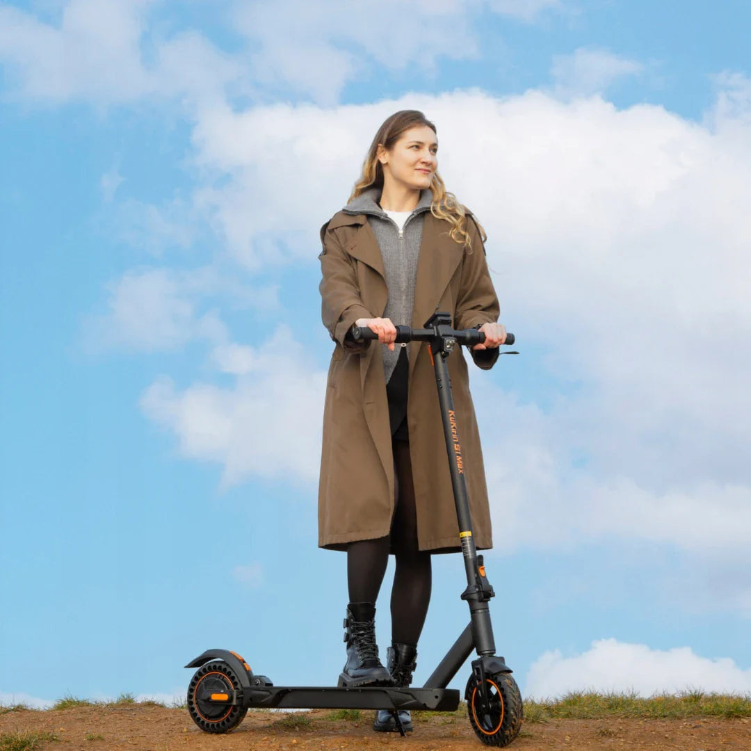 Izdelek na sliki je električni skiro, model Kukirin S1 Max. Na sliki je vidna ženska, ki stoji na skiroju v naravnem okolju, obdanem s svetlo modrim nebom in belimi oblaki. Oblečena je v dolgo jakno in črne škornje, kar daje vtis udobja in stila med vožnjo.
