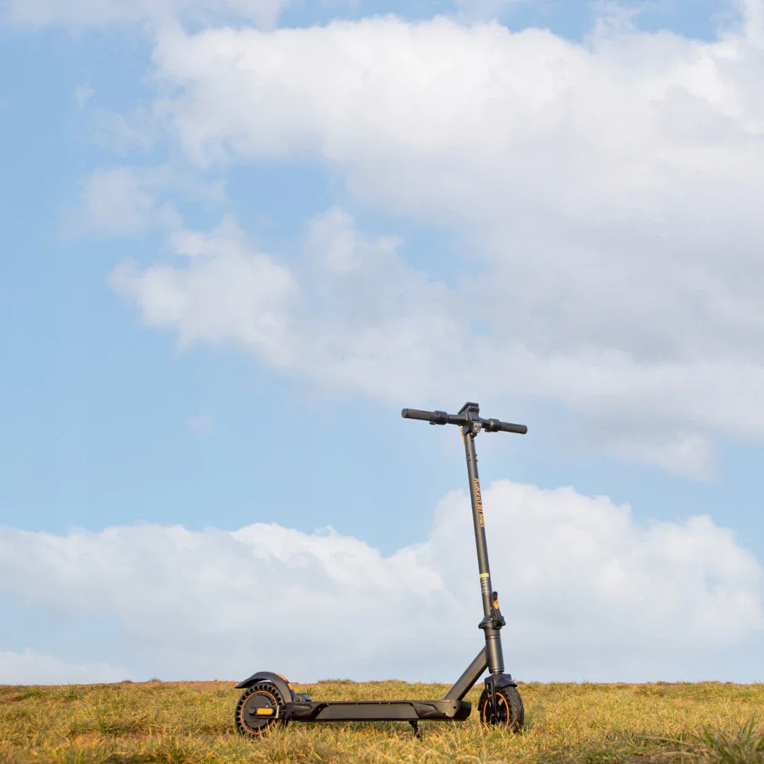 Na sliki je električni skiro znamke Kukirin, model S1 Max. Izdelek je postavljen na travnato površino, v ozadju pa je modro nebo s oblački. Skuter ima črno zasnovo s svetlimi detajli in je v pokončnem položaju, kar omogoča jasen pregled nad njegovim oblikovanjem.