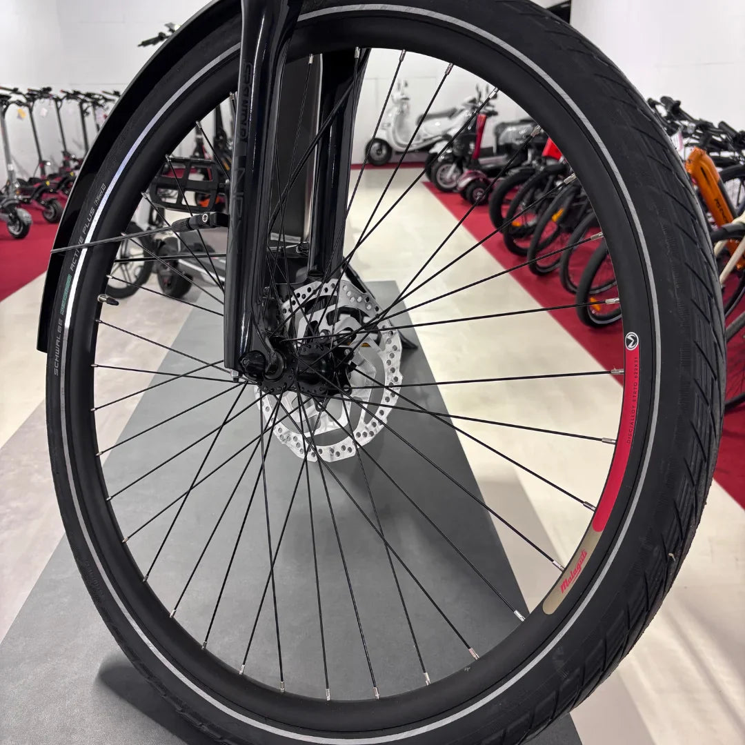 Izdelek na sliki je električno kolo znamke Malaguti, model Bolonina. Na sliki je prikazan sprednji kolo, ki vključuje disk zavore in pnevmatik z značilnim profilom. V ozadju so vidni tudi drugi električni skiroji in kolesa, kar nakazuje, da gre za prodajno mesto ali razstavo.