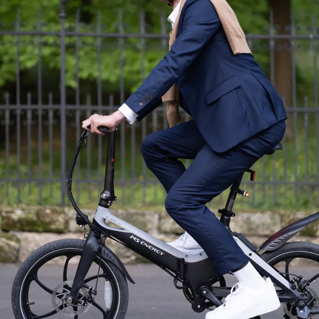 Na sliki je električno kolo znamke MS Energy, model i20. Oseba, oblečena v poslovno obleko, se vozi na kolesu, ki ima kompakten dizajn in manjše kolesa. Vidni so tudi elementi, kot so krmilo in sedež, kar nakazuje na udobno vožnjo. Okolje je zeleno, kar ustvarja prijetno vzdušje za vožnjo.