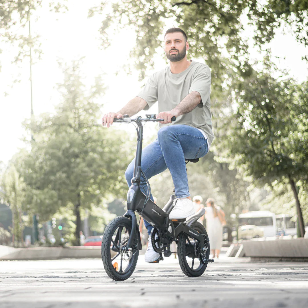 Na sliki je električno kolo, model MS Energy Urbanfold i6. Kolo je zložljivo in ima kompakten dizajn, kar ga naredi primernega za mestno vožnjo. Na sliki je moški, ki sedi na kolesu in se vozi po ulici, obdan z drevesi in mestno arhitekturo. Kolo je črne barve, opremljeno z manjšimi kolesi, kar dodatno poudarja njegovo praktičnost in mobilnost.