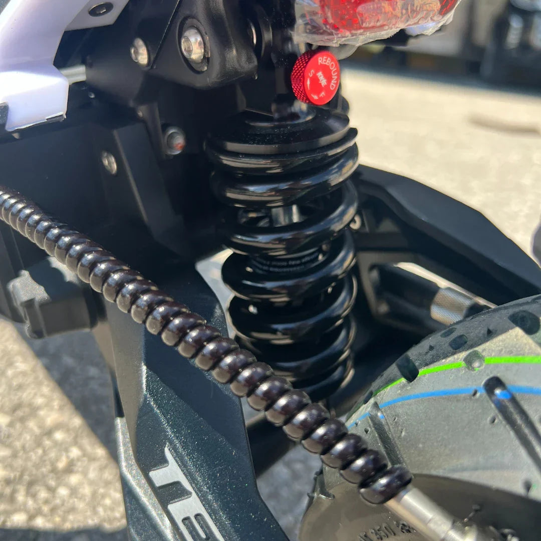 Izdelek na sliki je električni skiro, model Teverun Fighter Mini Pro 2025. Na sliki je prikazan del vzmetenja, ki vključuje črno vzmet ter del kolesa. Vidna je tudi struktura okvirja skiroja, ki je zasnovana za stabilnost in udobno vožnjo. Detajli, kot so vzmet in kolo, kažejo na robustno gradnjo, kar je značilno za to vrsto električnega prevoza.