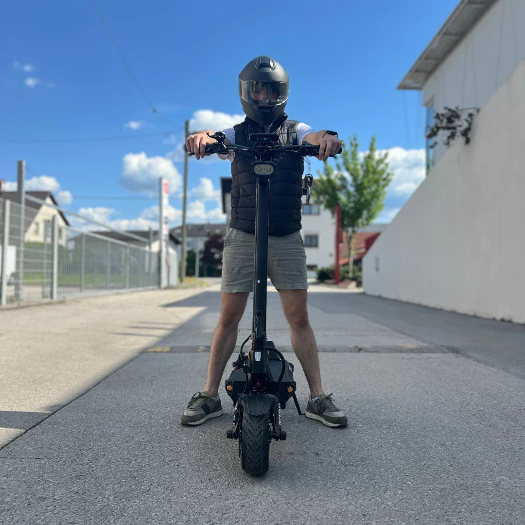 Na sliki je električni skiro znamke Teverun, model Fighter Supreme 7260 Ultra. Izdelek je prikazan z osebo, ki stoji na skiroju, oblečena je v zaščitno čelado in brezrokavnik. V ozadju je vidna cesta in nekaj stavb, kar nakazuje urbano okolje. Scooter ima robustno zasnovo in široke pnevmatike, kar daje vtis moči in stabilnosti.