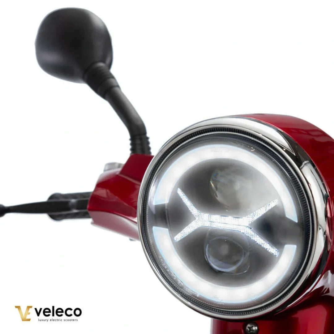 Izdelek je električni skuter, model Veleco Draco. Na sliki je prikazana sprednja luč skuterja, ki ima sodoben dizajn z LED tehnologijo. Vidni so tudi krmili in ogledalo, kar nakazuje na funkcionalnost in udobje pri vožnji. Skuter je v rdeči barvi, kar mu daje privlačen videz.