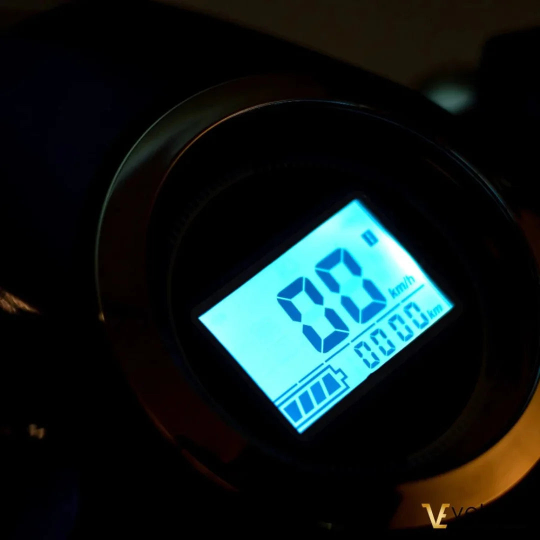 Izdelek je električni skiro znamke Veleco, model Faster. Na sliki je prikazan digitalni zaslon, ki prikazuje hitrost 0 km/h in prevoženo razdaljo 0,00 km. Zaslon vključuje tudi indikator napolnjenosti baterije, kar je pomembno za uporabnika.