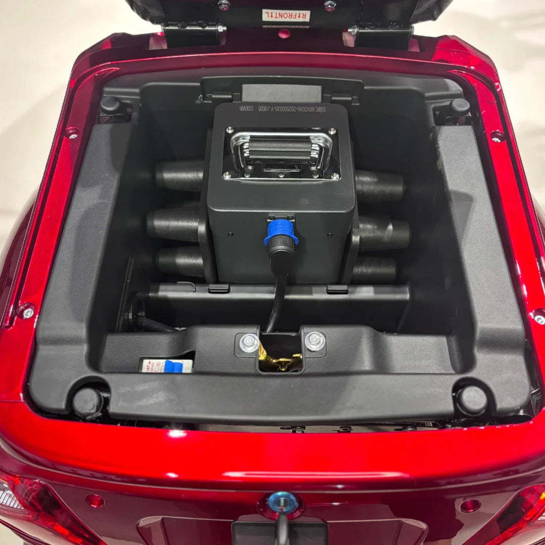 Izdelek je električni mobilni skuter s tremi kolesi, model Vigorous Vista 1. Na sliki je prikazan prostor za shranjevanje, ki se odpira, in v njem je vidna baterija ter električni sistem. Barva skuterja je rdeča, kar daje dinamičen videz.