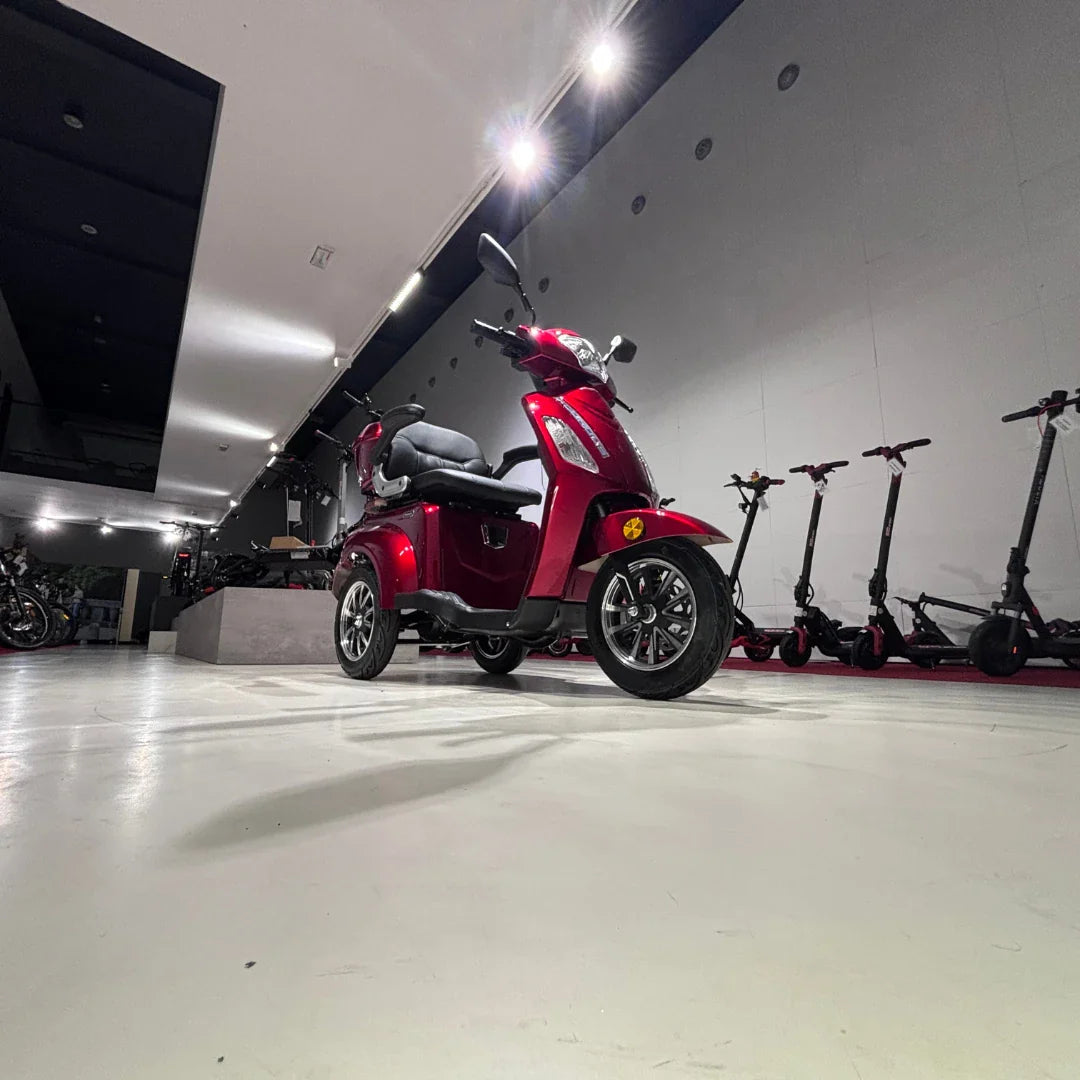 Izdelek na sliki je električni skuter s tremi kolesi, model Vigorous Vista 1. Skuter je rdeče barve in ima udobno sedežno površino ter elegantno obliko. V ozadju so vidni tudi električni skiroji, kar nakazuje, da je slika posneta v prodajnem prostoru za električna vozila.