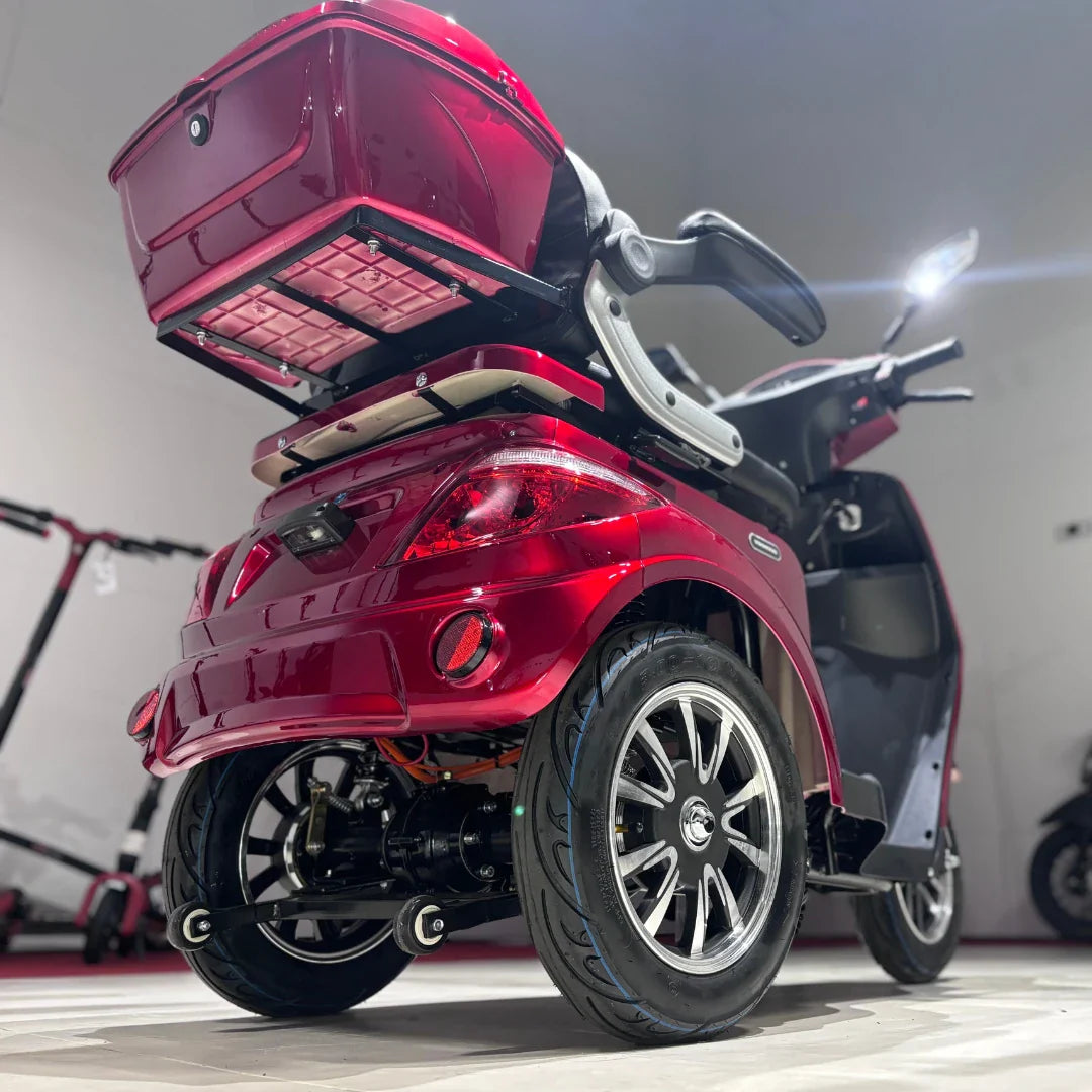 Izdelek na sliki je električni mobilni skuter z tremi kolesi, model Vigorous Vista 1. Značilna rdeča barva in oblikovanje nudita sodoben videz, medtem ko je na zadnjem delu skuterja vidna prtljažna enota, ki omogoča shranjevanje. Kolesa so široka, kar zagotavlja stabilnost, in skuter je opremljen s sodobnimi lučmi ter udobnim sedežem.