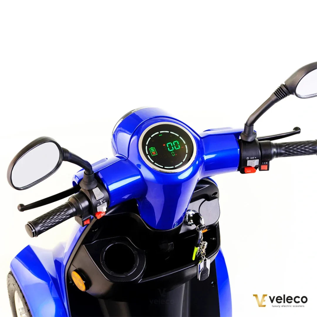 Na sliki je električni skiro znamke Veleco, model Gravis. Vidna je sprednja stran skiroja, kjer so prikazani krmili, merilnik hitrosti in nivoja baterije ter ogledali. Barva skiroja je modra, kar mu daje privlačen videz. Na krmilu so tudi različni gumbi za upravljanje.