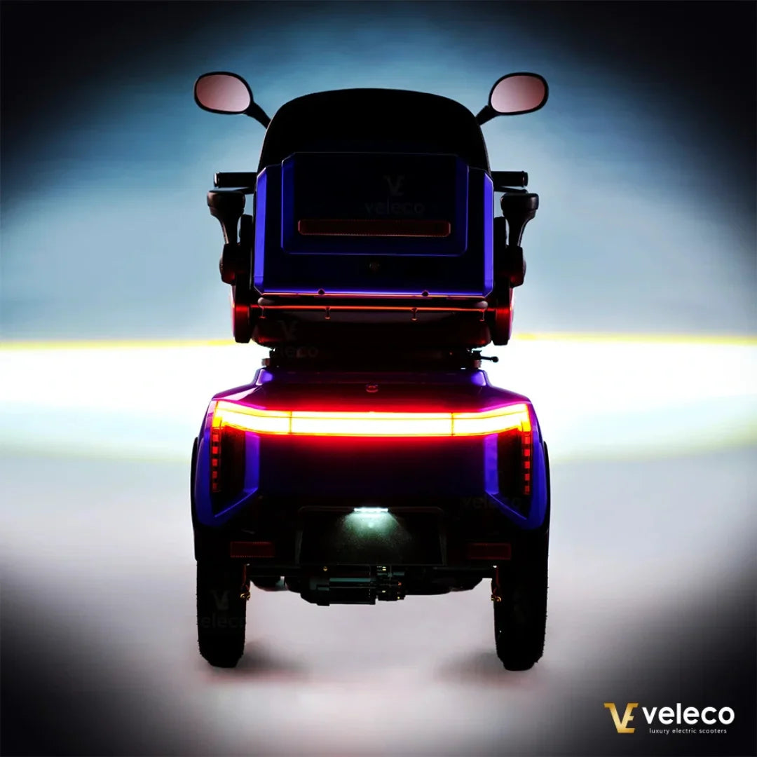 Izdelek na sliki je električni skuter, model Veleco Gravis. Na sliki je prikazan zadnji del skuterja, ki ima svetleče luči in značilno obliko. Skuter je v modri barvi in ima udoben sedež ter ogledala, kar nakazuje na njegovo uporabnost in udobje pri vožnji.