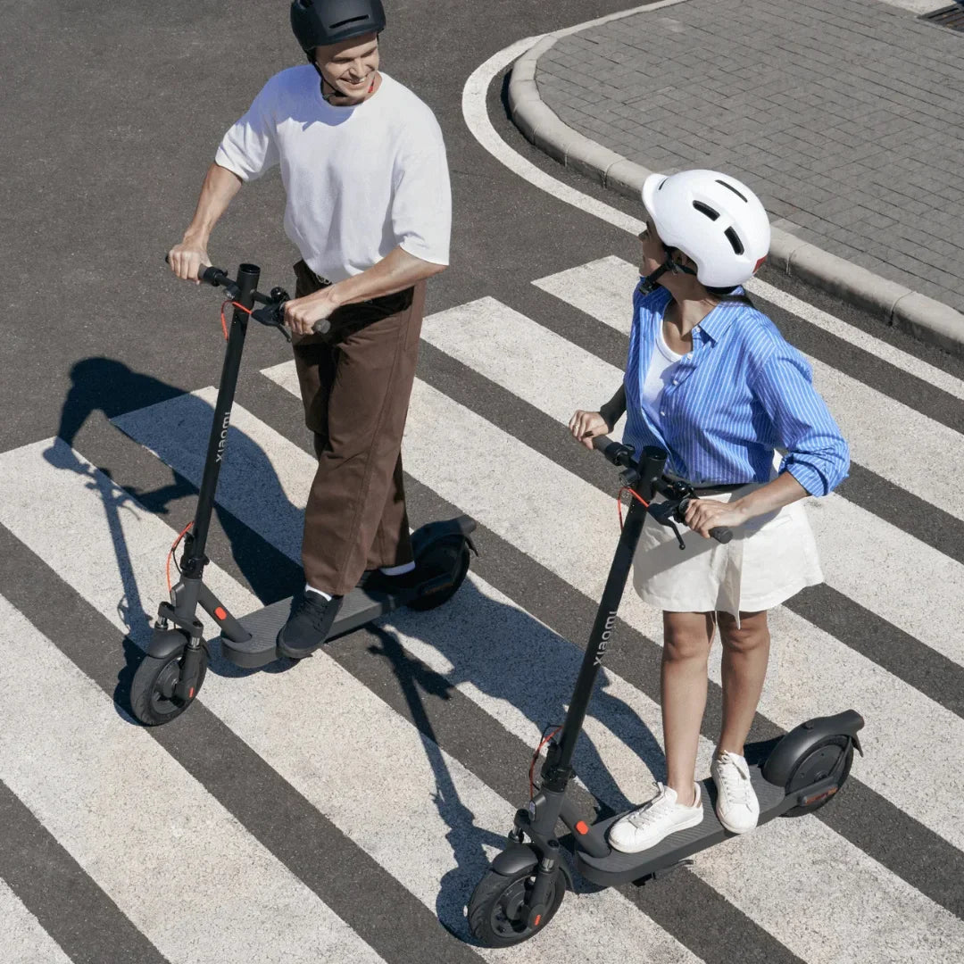 Na sliki je prikazan električni skiro znamke Xiaomi, model Elite. Dva uporabnika, moški in ženska, se peljeta po prehodu za pešce. Oba nosita čelade za zaščito, kar nakazuje na varno vožnjo. Ozadje vključuje asfaltirano cesto in peščen prehod, kar ustvarja urbano okolje.