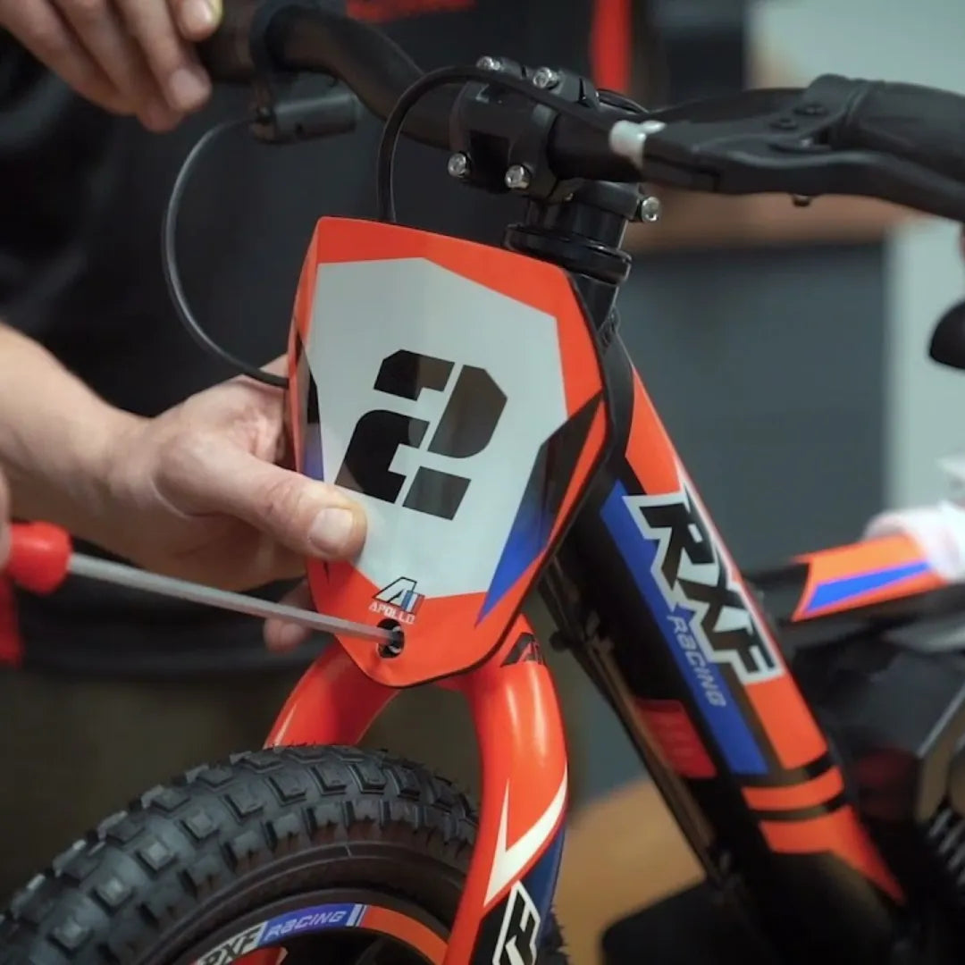 Izdelek na sliki je električno kolo, model Apollo Sedna 12 RXF. Na sliki je vidna oseba, ki drži kolo in uporablja orodje za prilagajanje dela kolesa. Kolo ima živahno barvno zasnovo v rdeči, modri in črni barvi, z značilnimi oznakami RXF in logotipom znamke Apollo. Vidna je tudi pnevmatika, ki je primerna za različne terene.