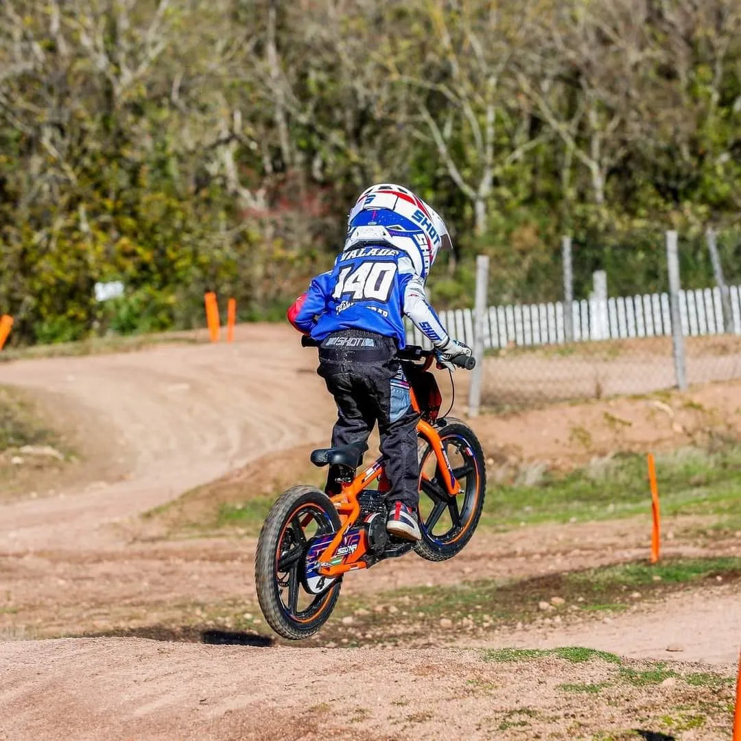 Izdelek je električno kolo, model Apollo Sedna 16 RXF. Na sliki je otrok, ki vozi kolo po terenski stezi, medtem ko skakanje z dvignjenim kolesom. Kolo je živih barv, predvsem oranžne, in je zasnovano za vožnjo po razgibanih terenih. V ozadju so vidne naravne površine in ograja, kar nakazuje, da gre za kolesarsko progo.