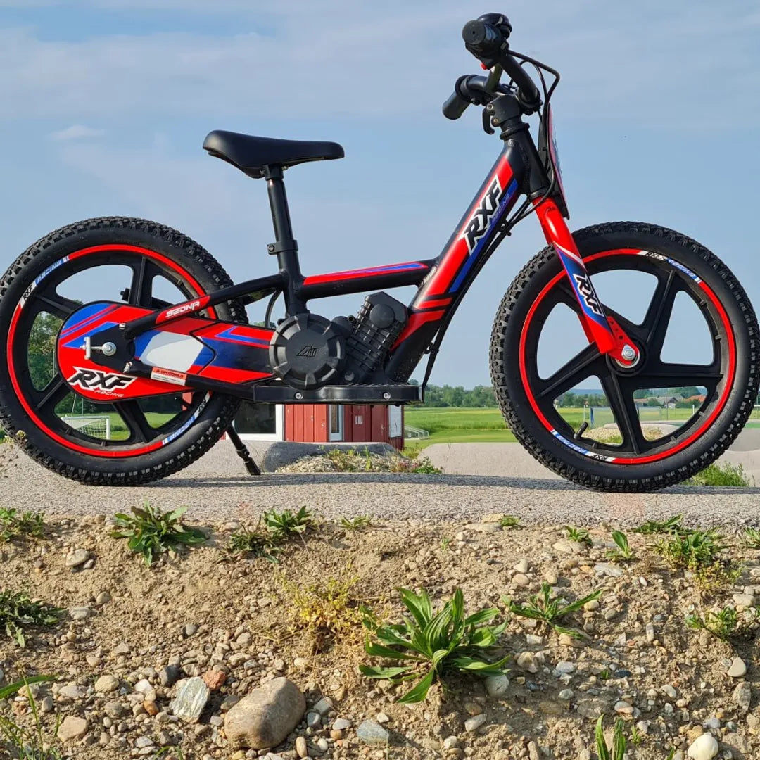 Izdelek na sliki je električno kolo, znamke RXF, model Sedna 16 RXF. Na sliki je vidno kolo z rdeče-modro barvno kombinacijo, širokimi gumami in športnim dizajnom. Kolo ima robusten okvir in je postavljeno na asfaltno površino, v ozadju pa je vidna zelena pokrajina.