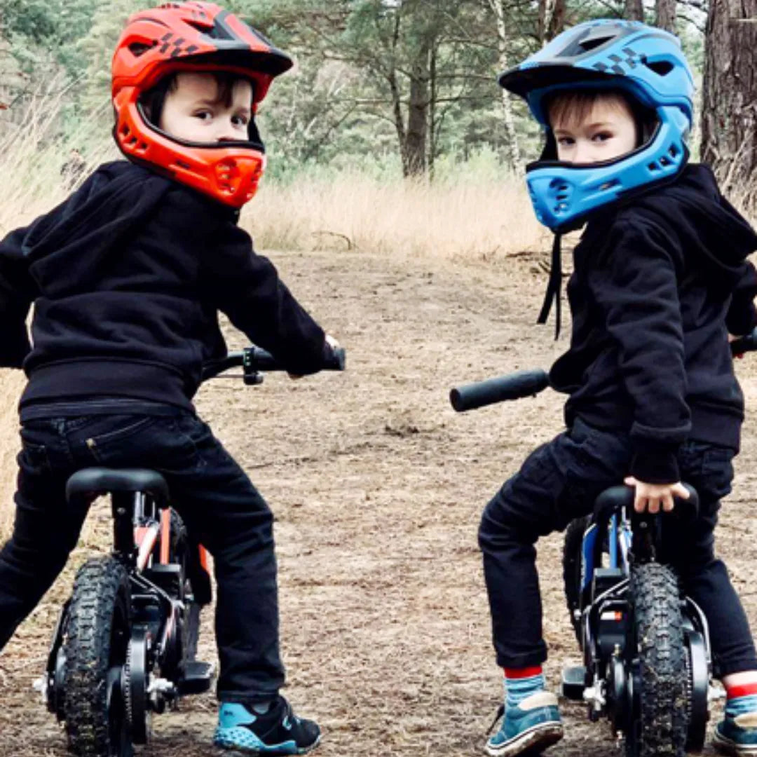 Izdelek je električno kolo, model Apollo Sedna 16 RXF. Na sliki sta vidna dva otroka, ki sedita na svojih kolesih, oblečena v črna oblačila in zaščitne čelade, ena rdeča in ena modra. Okolica je gozdnata, kar nakazuje, da se kolesarita po naravni poti.