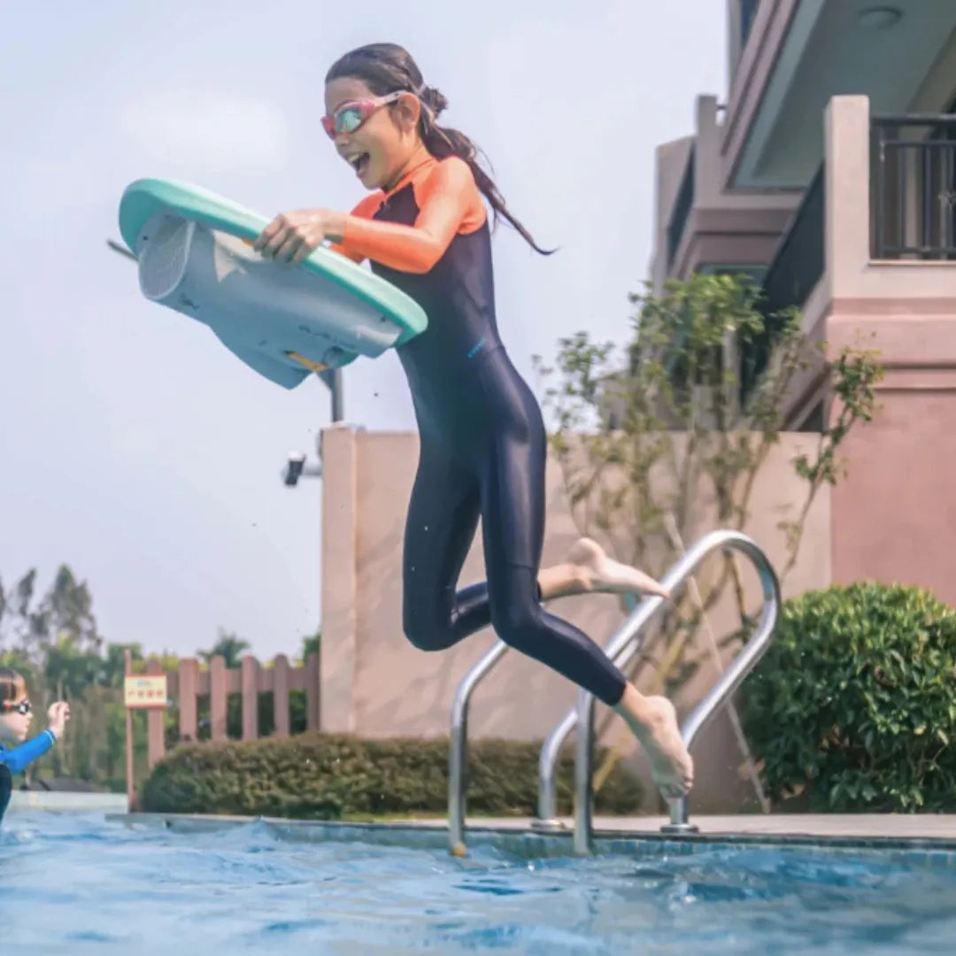 Izdelek je električna plavalna deska znamke Asiwo, model Mako. Na sliki je vidna deklica v neoprenu, ki skoči v bazen z električno plavalno desko v rokah. V ozadju je bazenska ograja in zelenje, kar ustvarja sproščeno poletno vzdušje.