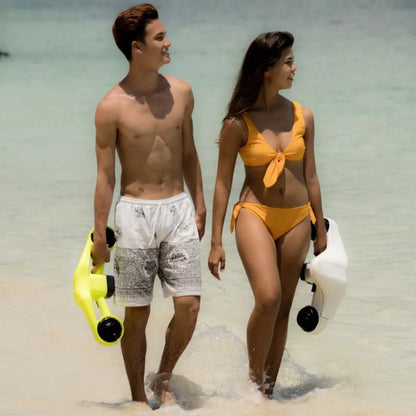 Izdelek je električni podvodni skuter znamke Asiwo, model Manta. Na sliki sta dva mlada človeka, ki hodita po obali, oblečena v kopalne obleke. Vsak drži enega izmed skuterjev, enega rumenega in enega belega, kar nakazuje na aktivno preživljanje časa v vodi. Obe osebi se smejeta in uživata v sončnem vremenu.