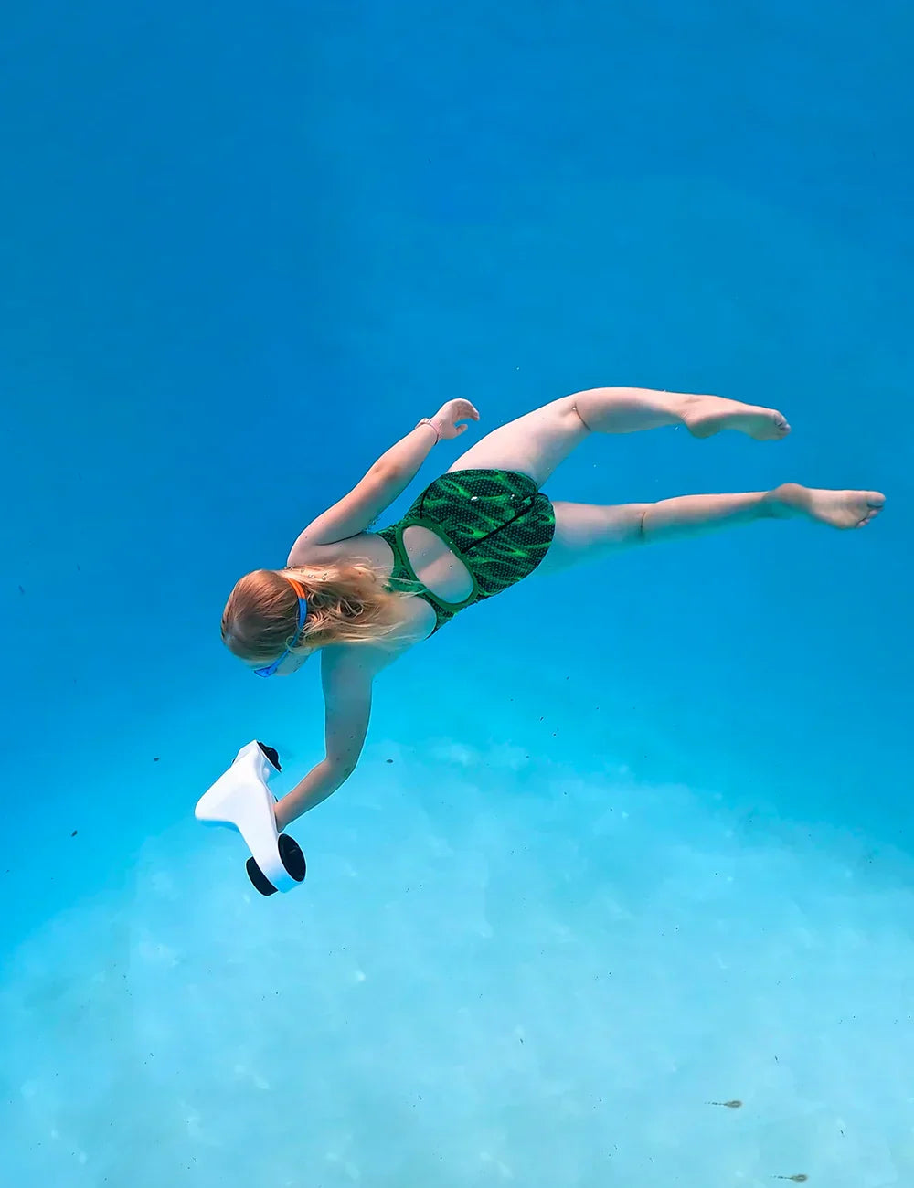 Izdelek na sliki je električni podvodni skuter, model Asiwow Manta. Na sliki je vidna deklica, ki plava pod vodo in drži skuter, oblečena v kopalke z zelenim vzorcem. V ozadju je jasno modro vodo, kar ustvarja prijetno podvodno vzdušje.