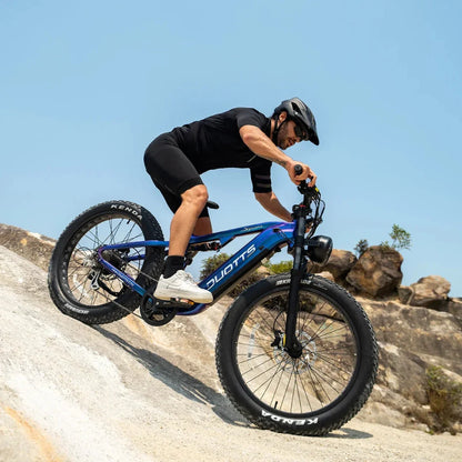 Na sliki je električno kolo znamke Duotts, model E26. Kolo ima široke gume, kar omogoča boljšo stabilnost in oprijem na različnih terenih. Na sliki je vidna oseba, ki vozi kolo po skalnati površini, oblečena je v športno opremo in čelado, kar kaže na aktivno in avanturistično uporabo kolesa. Ozadje je jasno modro nebo, kar dodatno poudarja dinamičnost prizora.