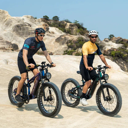 Na sliki sta prikazana dva moška, ki vozita električna kolesa. Izdelek je električno kolo, model E26, znamke Duotts. Obe kolesi imata široke pnevmatike, kar omogoča vožnjo po različnih terenih, in sta zasnovani za aktivno uporabo na prostem. Kolesarji nosijo športna oblačila in čelade, kar nakazuje na športno naravo dejavnosti. V ozadju je naravna pokrajina z skalami in zelenjem.