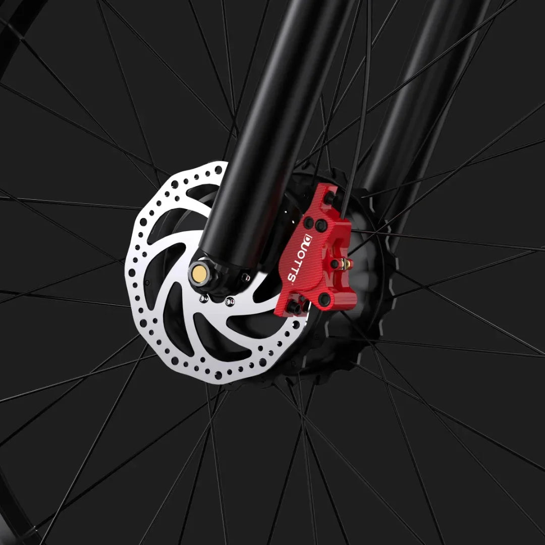 Izdelek je električno kolo, znamke Duotts, model F26. Na sliki je prikazan del sprednjega kolesa, kjer je vidna disk zavora s kovinsko ploščo in rdečim zavorni kaliperjem. Kolo ima temno barvo, kar daje sodoben in športen videz.