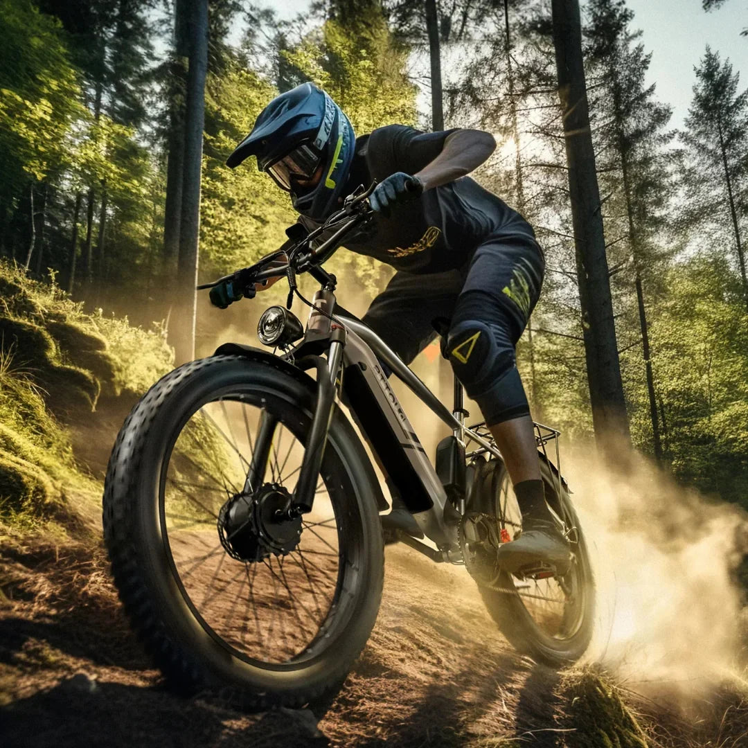 Izdelek na sliki je električno kolo, model Duotts F26. Na sliki je kolesar, oblečen v zaščitno opremo, ki vozi po gozdni poti, obdan z naravo. Kolo ima široke pnevmatike, kar nakazuje na njegovo zmogljivost na terenu, in je opremljeno z lučjo spredaj. Kolesar se zdi osredotočen in uživa v vožnji po prašni poti.