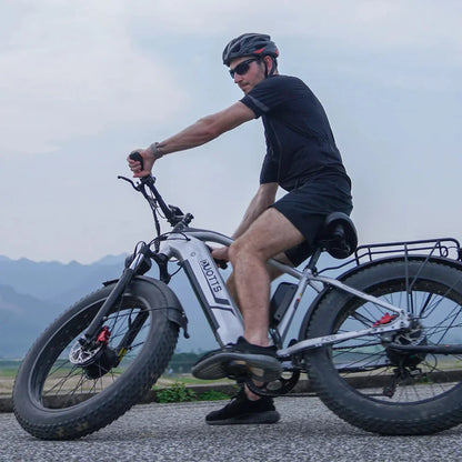 Na sliki je električno kolo znamke DUOTTS, model F26. Kolo ima široke gume, kar omogoča boljšo stabilnost in vožnjo po različnih terenih. Moški, oblečen v športna oblačila in čelado, se vozi po cesti, v ozadju pa so vidne gore in narava.