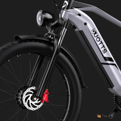 Izdelek na sliki je električno kolo znamke DUOTTS, model F26. Na sliki je prikazan sprednji del kolesa, ki vključuje široko pnevmatiko, sprednje vzmetenje in disk zavore. Vidna je tudi sprednja luč, kar nakazuje na uporabo kolesa v različnih svetlobnih pogojih. Kolo ima sodoben in robusten dizajn, kar ga naredi primernega za različne terene.