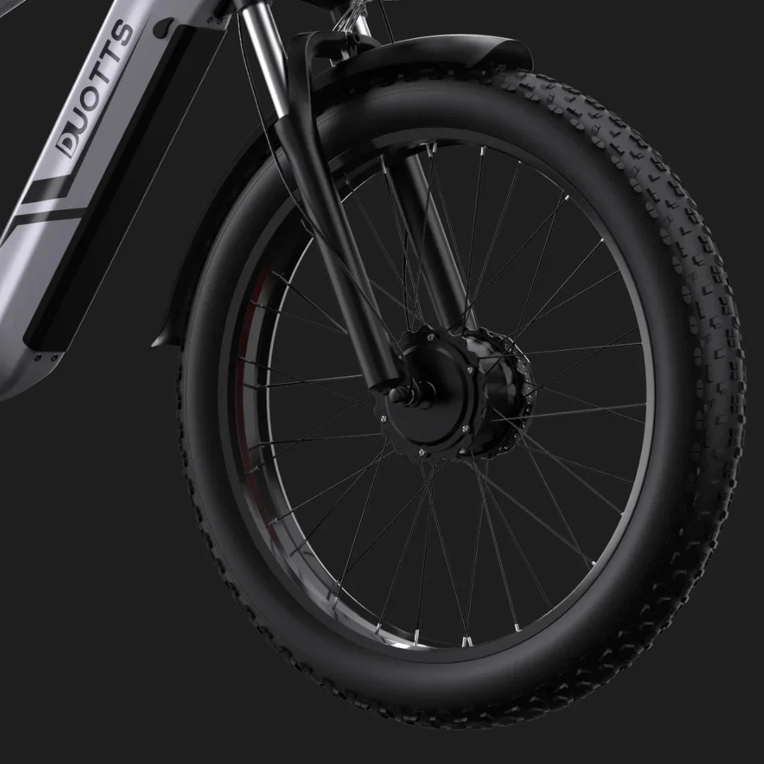 Izdelek na sliki je električno kolo, znamke DUOTTS, model F26. Na sliki je prikazan sprednji del kolesa, ki vključuje široko pnevmatiko z izrazitim profilom, kar nakazuje na primernost za različne terene. Vidna je tudi sprednja vilica in motor v osi kolesa, kar je značilno za električna kolesa. Celotna zasnova je moderna in robustna.