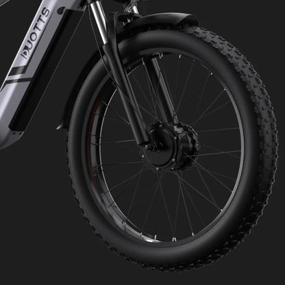 Izdelek na sliki je električno kolo, znamke DUOTTS, model F26. Na sliki je prikazan sprednji del kolesa, ki vključuje široko pnevmatiko z izrazitim profilom, kar nakazuje na primernost za različne terene. Vidna je tudi sprednja vilica in motor v osi kolesa, kar je značilno za električna kolesa. Celotna zasnova je moderna in robustna.