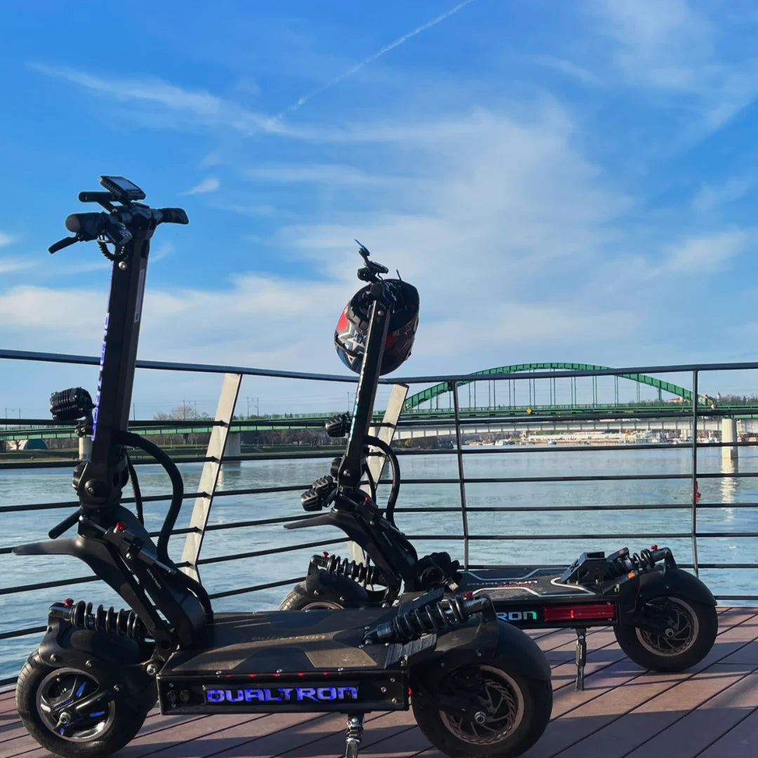Dva električna skiroja Dualtron X Limited parkirana ob reki z mostom v ozadju.