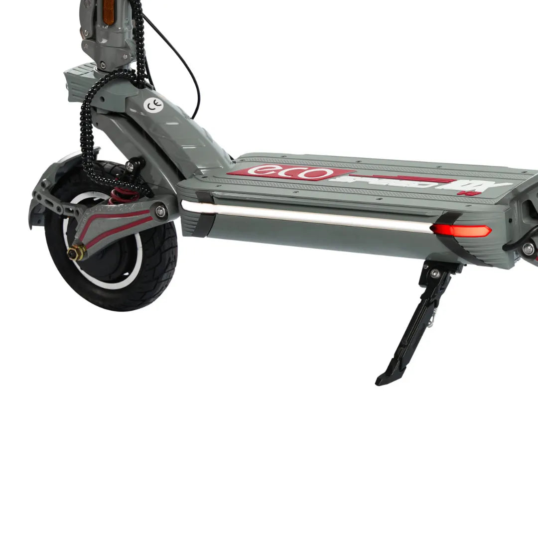 Izdelek je električni skiro, model Eco Speed 10X V2. Na sliki je vidna zadnja kolesna enota skiroja, ki vključuje široko kolo z belo obrobo in vzmetenje. Vidna je tudi platforma za stojišče, ki je opremljena z oznako znamke. Dodatno je prisoten tudi nosilec za stojalo, kar omogoča stabilno parkiranje skiroja. Celoten dizajn je sodoben in funkcionalen.