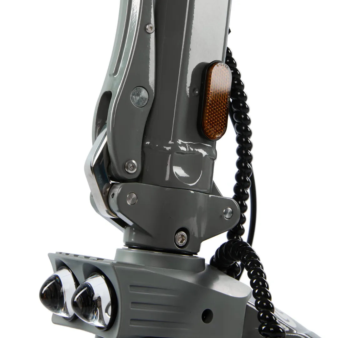 Izdelek je električni skiro, model Eco Speed 10X V2. Na sliki je prikazan del skiroja, ki vključuje mehanizem za zlaganje, svetlobno oznako v oranžni barvi ter dva sprednja reflektorja. Barva skiroja je siva, kar mu daje sodoben videz. Vidna je tudi spiralna zaščita kabla, kar kaže na premišljen dizajn in varnost.