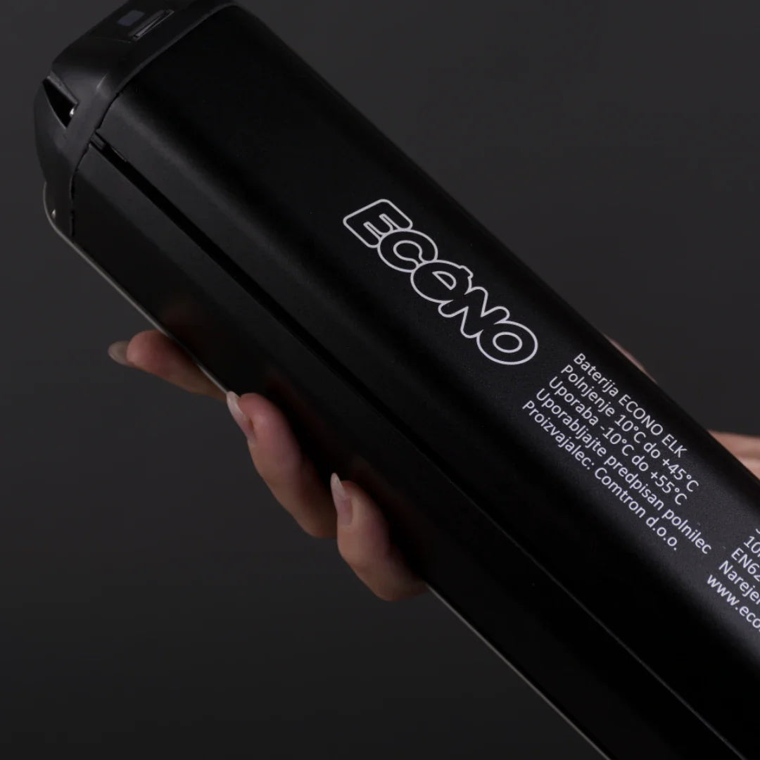 Na sliki je vidna baterija za električno kolo znamke Econo, model ELC4. Baterija je črne barve in ima jasno označeno ime znamke ter specifikacije, kot so temperaturne meje delovanja in navodila za uporabo. Oseba v sliki drži baterijo, kar nakazuje, da je izdelek prenosljiv in enostaven za uporabo.