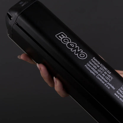 Na sliki je vidna baterija za električno kolo znamke Econo, model ELC4. Baterija je črne barve in ima jasno označeno ime znamke ter specifikacije, kot so temperaturne meje delovanja in navodila za uporabo. Oseba v sliki drži baterijo, kar nakazuje, da je izdelek prenosljiv in enostaven za uporabo.